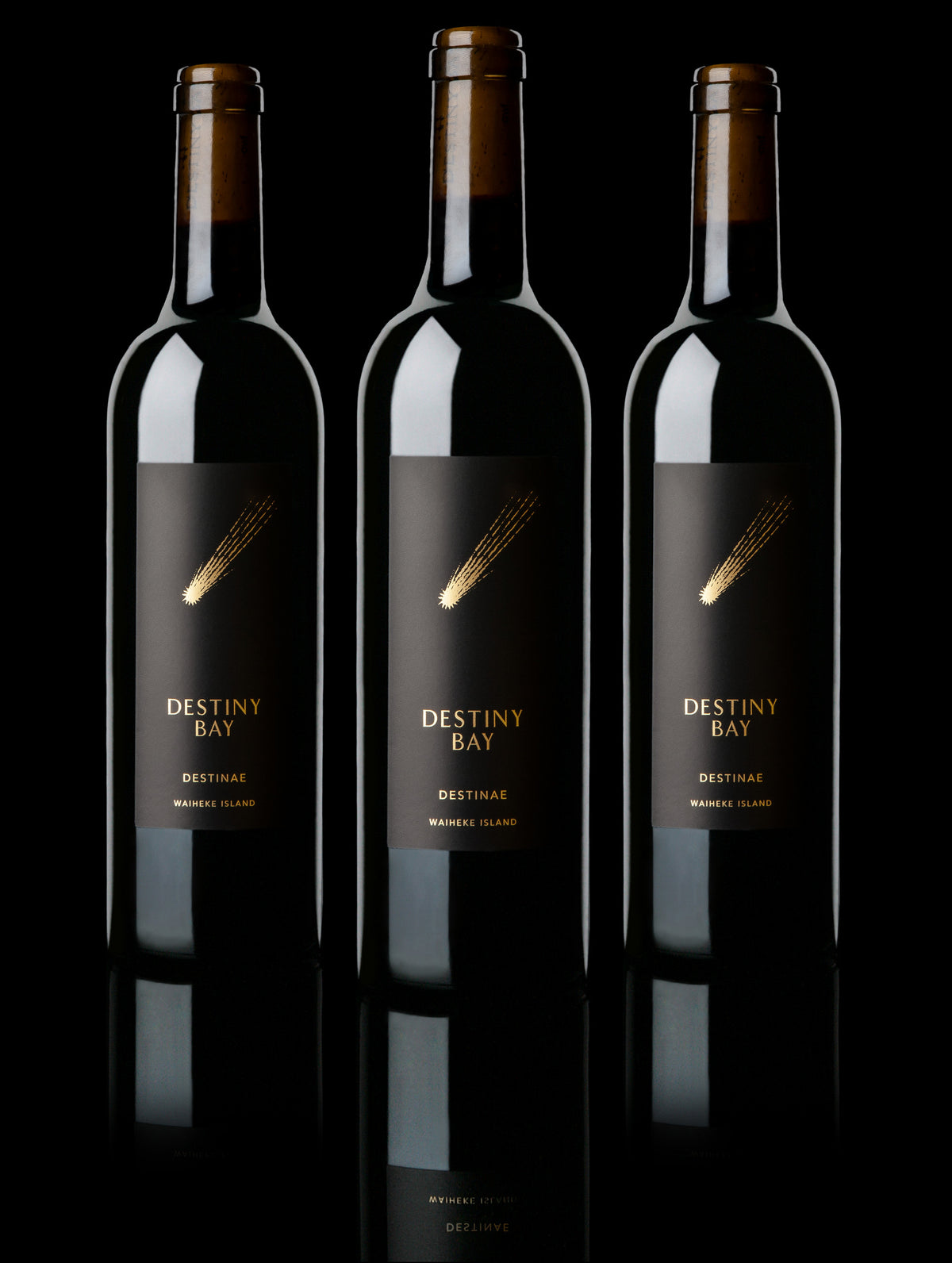 Three dark glass wine bottles with black labels and gold 'DESTINY BAY' branding, arranged side by side on a black reflective surface against a black background.
